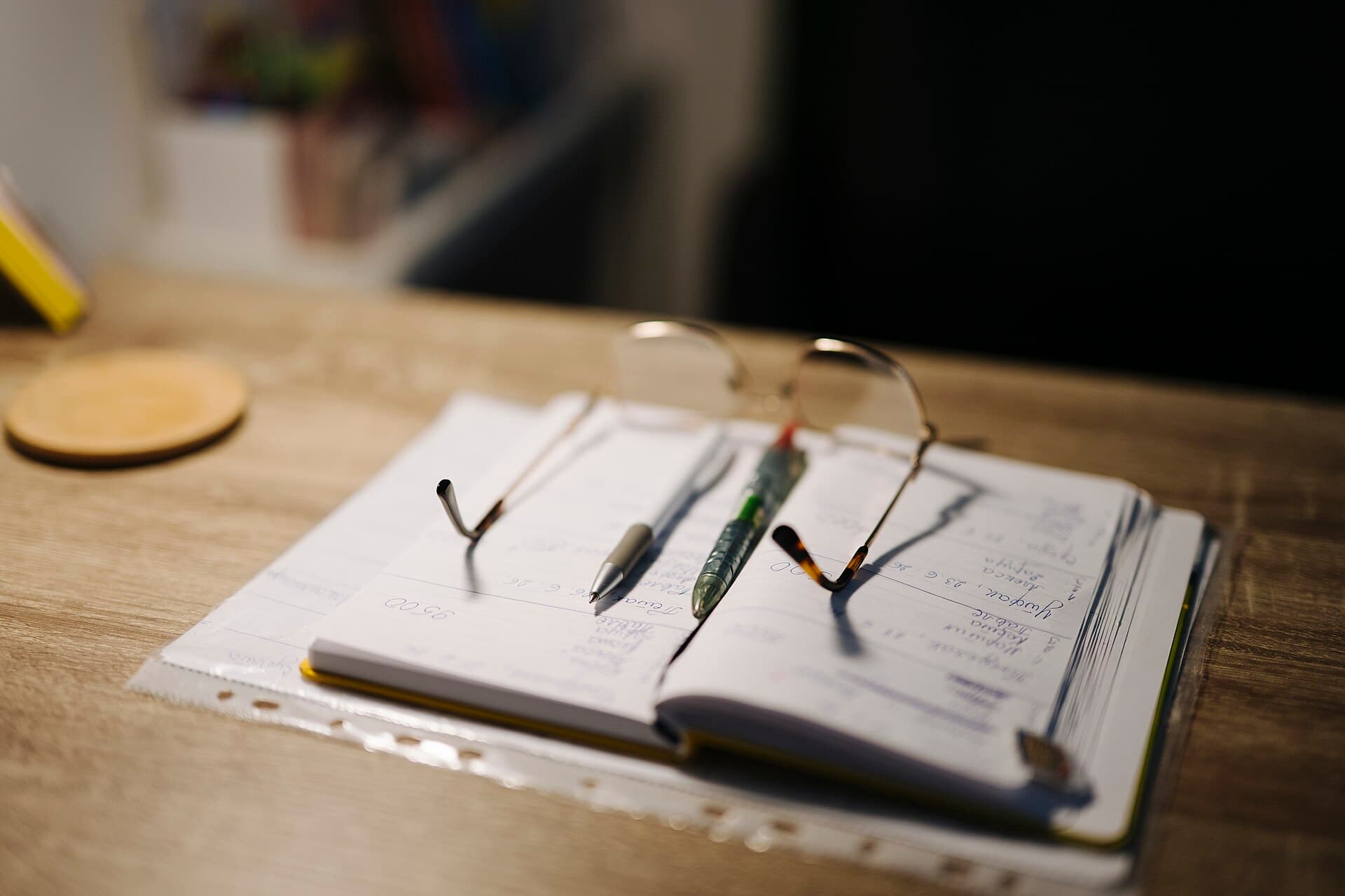 An open notebook, pens and glasses on a desk
